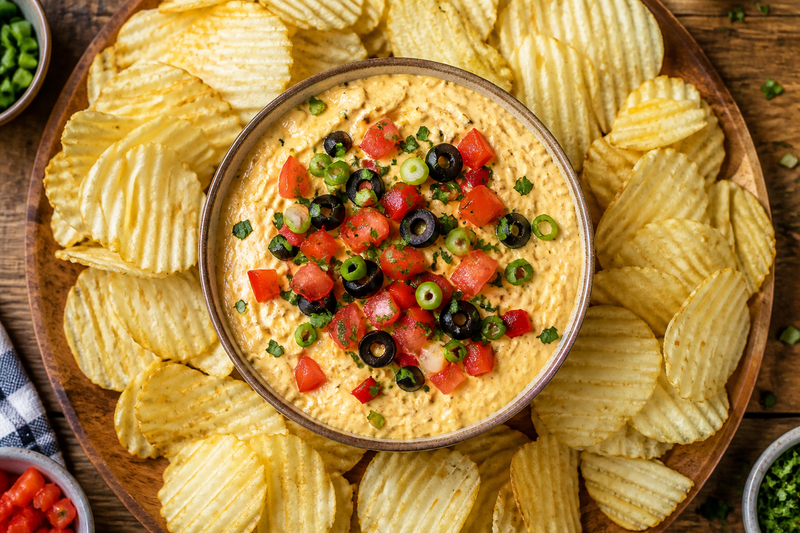 A bowl of whipped pub cheese topped with diced vegetables, surrounded by RUFFLES potato chips for dipping